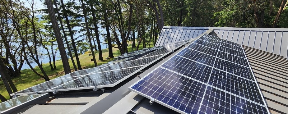 Rooftop view of solar panels on the roof of an off grid home. The ocean is visible through the trees in the background.