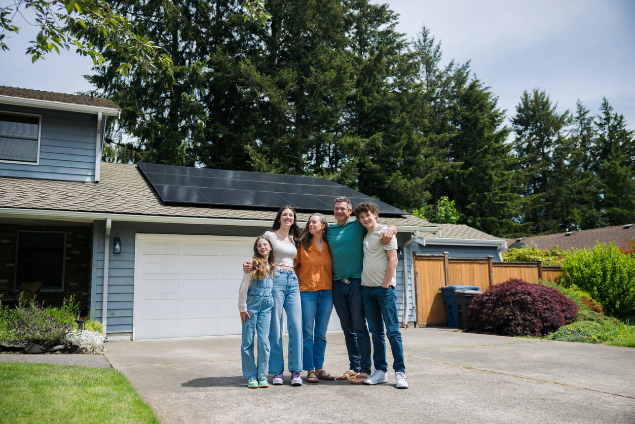 A family of 5 standing in their driveway, with their solar panels in view on their garage roof in the background.