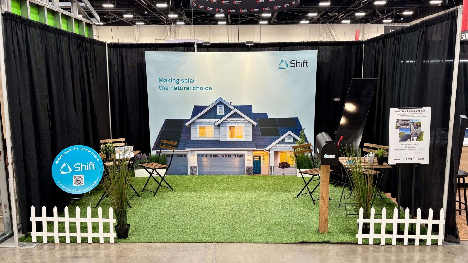 A booth at the Calgary Home Show. It is decorated to look like the front lawn of someone's home. There is a mail box, artificial turf, lawn furniture, a fenceline, and a large banner at the back of a home with solar panels on it.