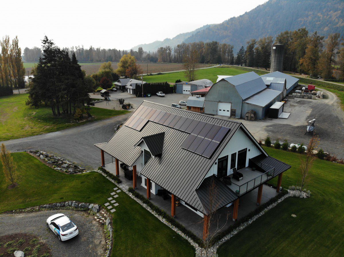 aerial view of a modern home in a rural farm setting. There are solar panels on the metal roof of the home, and there are barns and farm buildings in the background.